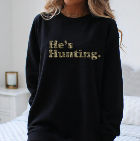 Black sweatshirt with 'He's Hunting' text in gold on a neutral background