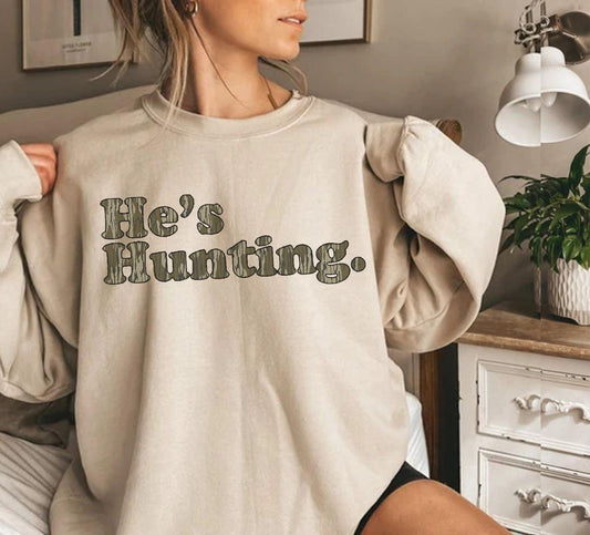 Person wearing a beige sweatshirt with 'He's Hunting.' text in a home setting.