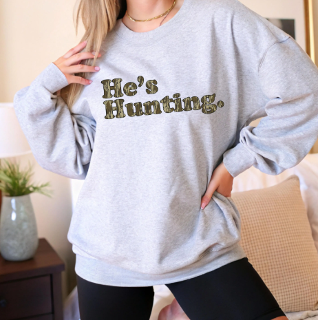 Person wearing a gray sweatshirt with 'He's Hunting.' text in a home setting