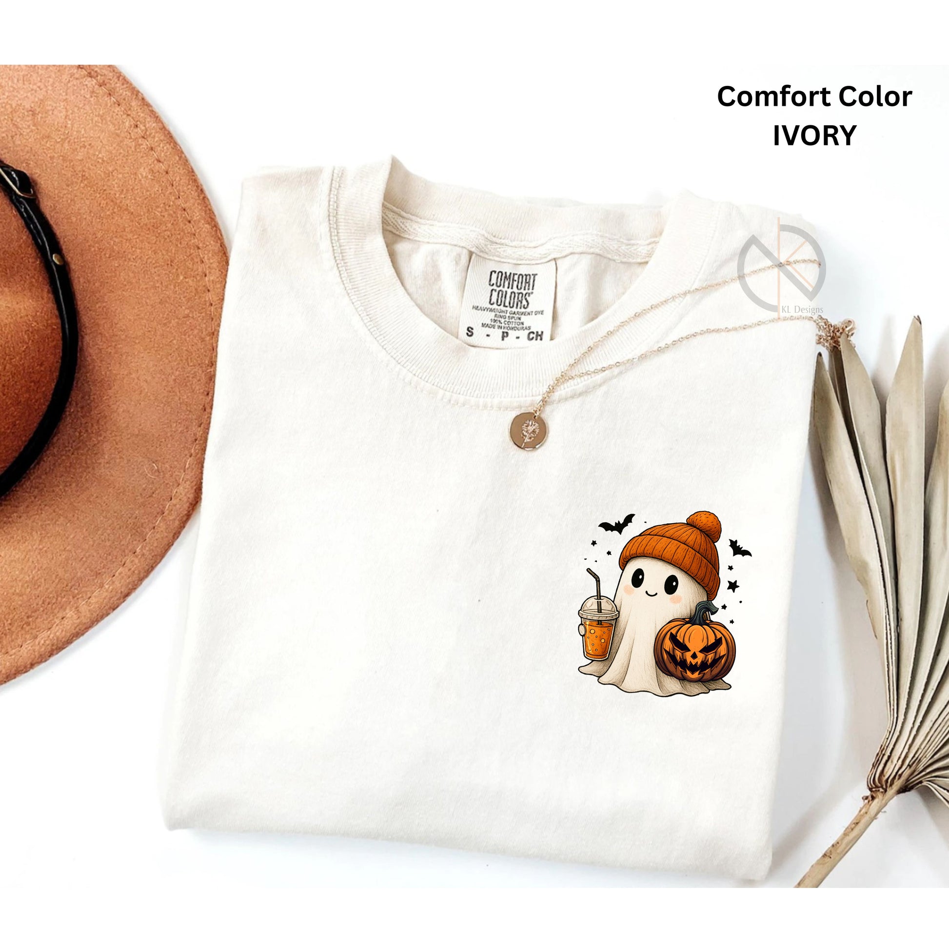 White t-shirt with a ghost and pumpkin design, placed on a white surface with a hat and dried leaves.