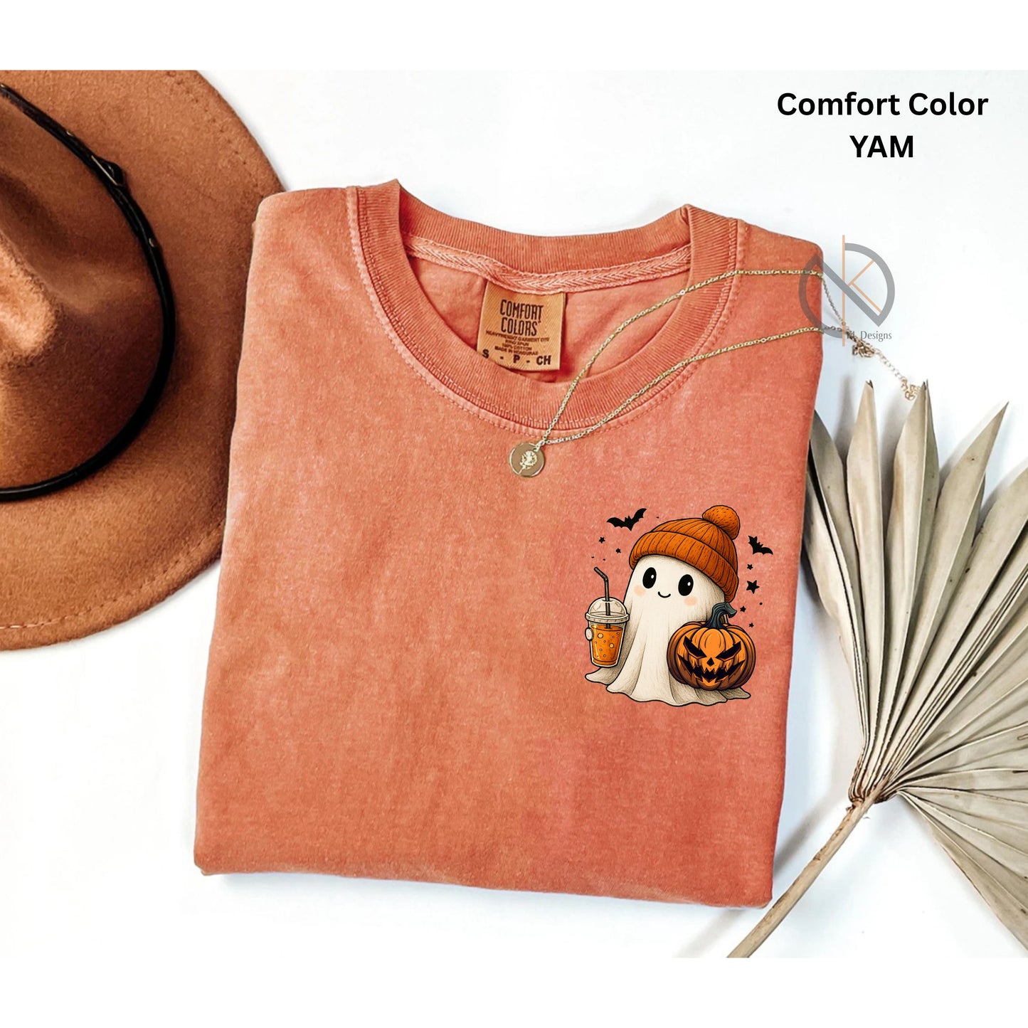 Folded yam-colored t-shirt with ghost and pumpkin design, next to a brown hat and dried leaves.